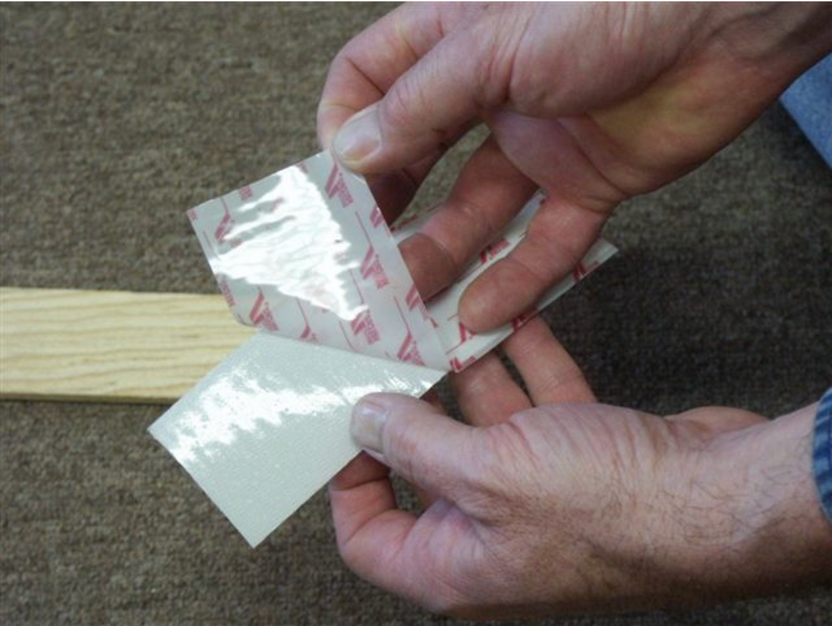 Hands peeling the paper backing off self-adhesive Velcro tape beside a thin wood strip